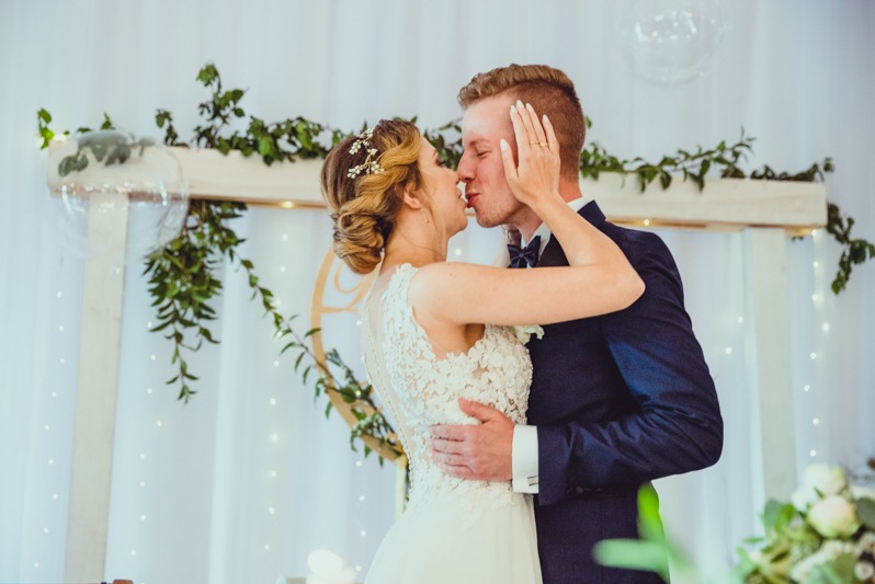 Pocałunek pary młodej na tle drewnianego łuku z zielenią i białymi zasłonami z drobnymi światełkami. Pan młody w granatowym garniturze, panna młoda w białej sukni.