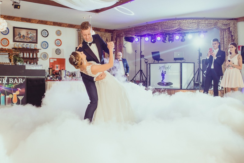 Para młoda tańczy w chmurach dymu na sali weselnej. W tle DJ, goście i dekoracje. Ujęcie z niskiego kąta, podkreślające taniec i atmosferę zabawy.