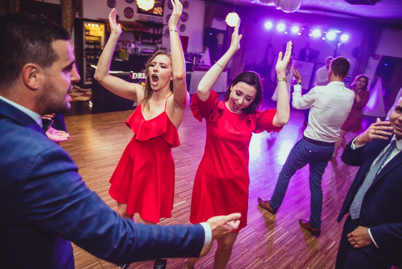 Rozbawieni goście w czerwonych sukienkach tańczą na parkiecie podczas imprezy. W tle oświetlenie sceniczne i bar. Dynamiczne ujęcie z perspektywy uczestnika zabawy.