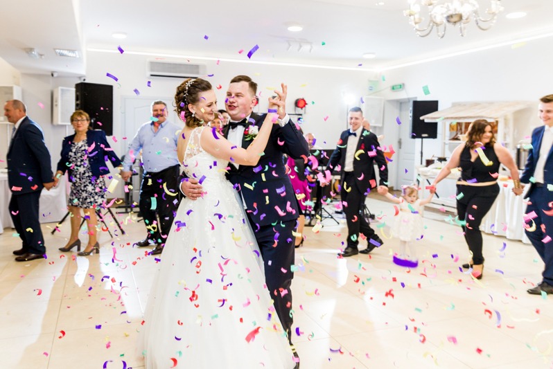 Para młoda w tańcu podczas wesela, obsypana konfetti, w tle goście trzymający się za ręce. Jasne wnętrze sali z żyrandolem i elementami dekoracyjnymi.