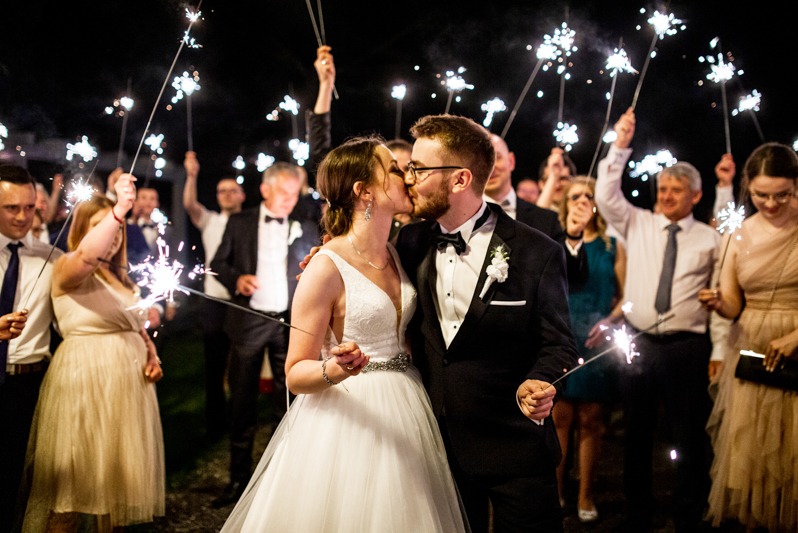 Pocałunek nowożeńców otoczonych gośćmi z zimnymi ogniami podczas wesela w nocy. Panna młoda w sukni z kryształowym pasem, pan młody w smokingu.