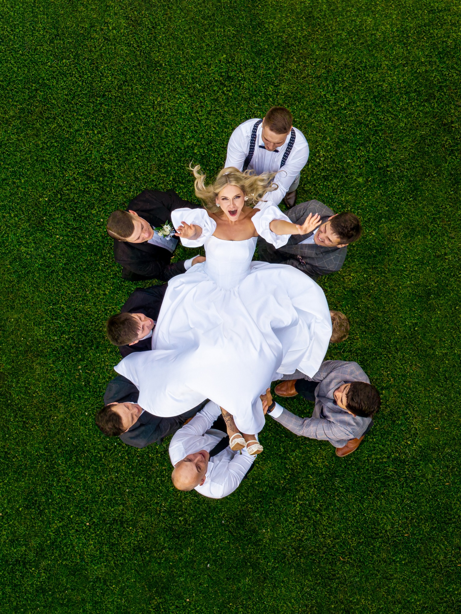 Panna młoda w białej sukni unoszona przez drużbów na tle zielonej trawy, widok z góry, dynamiczne ujęcie.