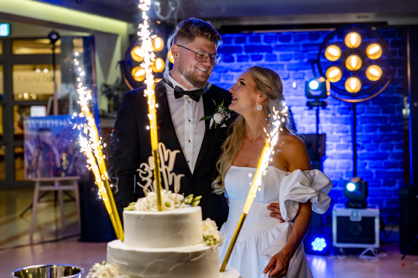 Uśmiechnięta para młoda przy torcie weselnym z fontannami iskier na tle niebieskiej ściany z dekoracyjnym oświetleniem.