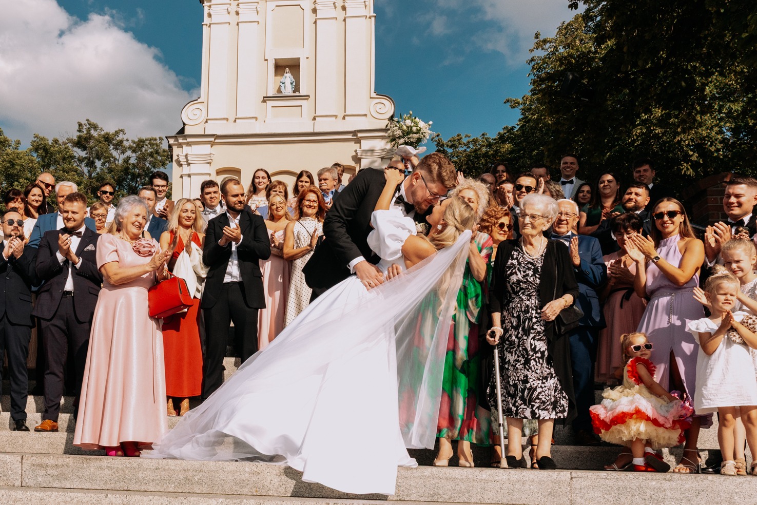 Pocałunek pary młodej na schodach kościoła, otoczonej przez gości weselnych. Panna młoda w białej sukni, pan młody w czarnym garniturze. Słoneczny dzień, radosna atmosfera.