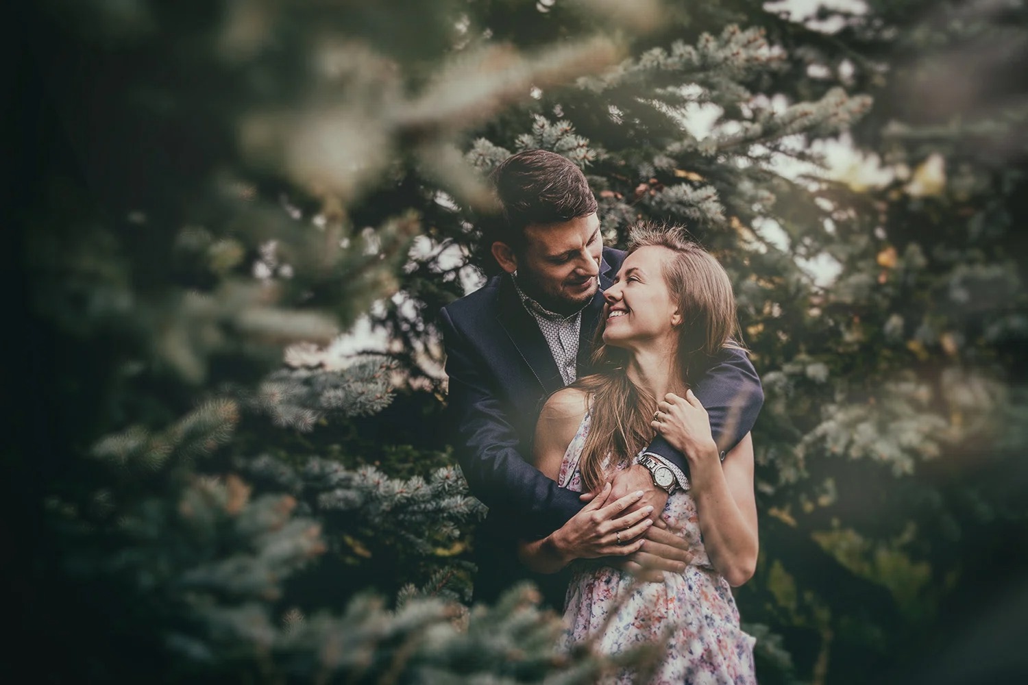 Uśmiechnięta para w objęciach na tle zielonych drzew iglastych. Kobieta w kwiecistej sukience, mężczyzna w marynarce. Romantyczna atmosfera, naturalne światło.