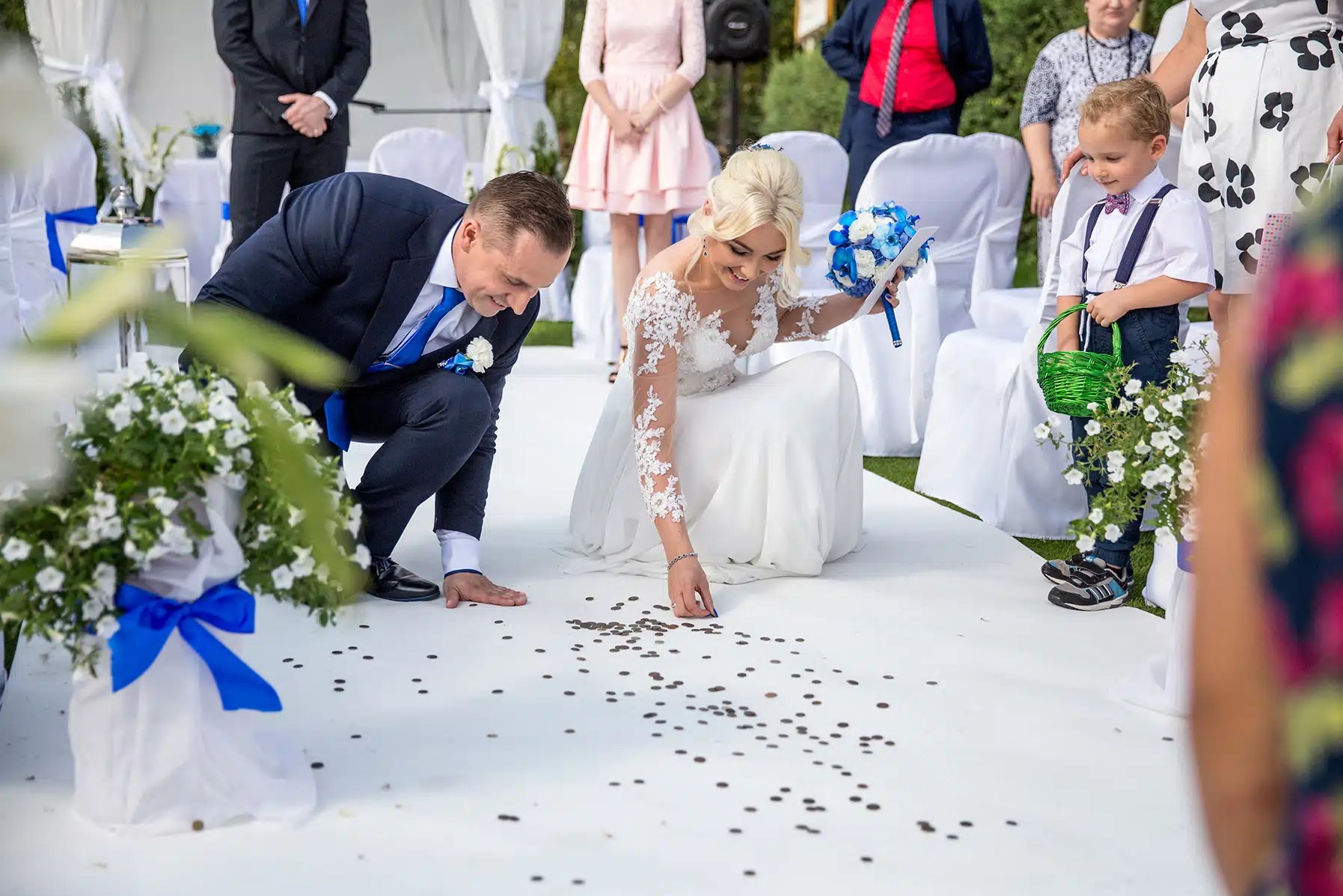 Para młoda zbiera monety na białym dywanie podczas ceremonii ślubnej w plenerze. Widoczny bukiet panny młodej i kwiaty z niebieską wstążką.