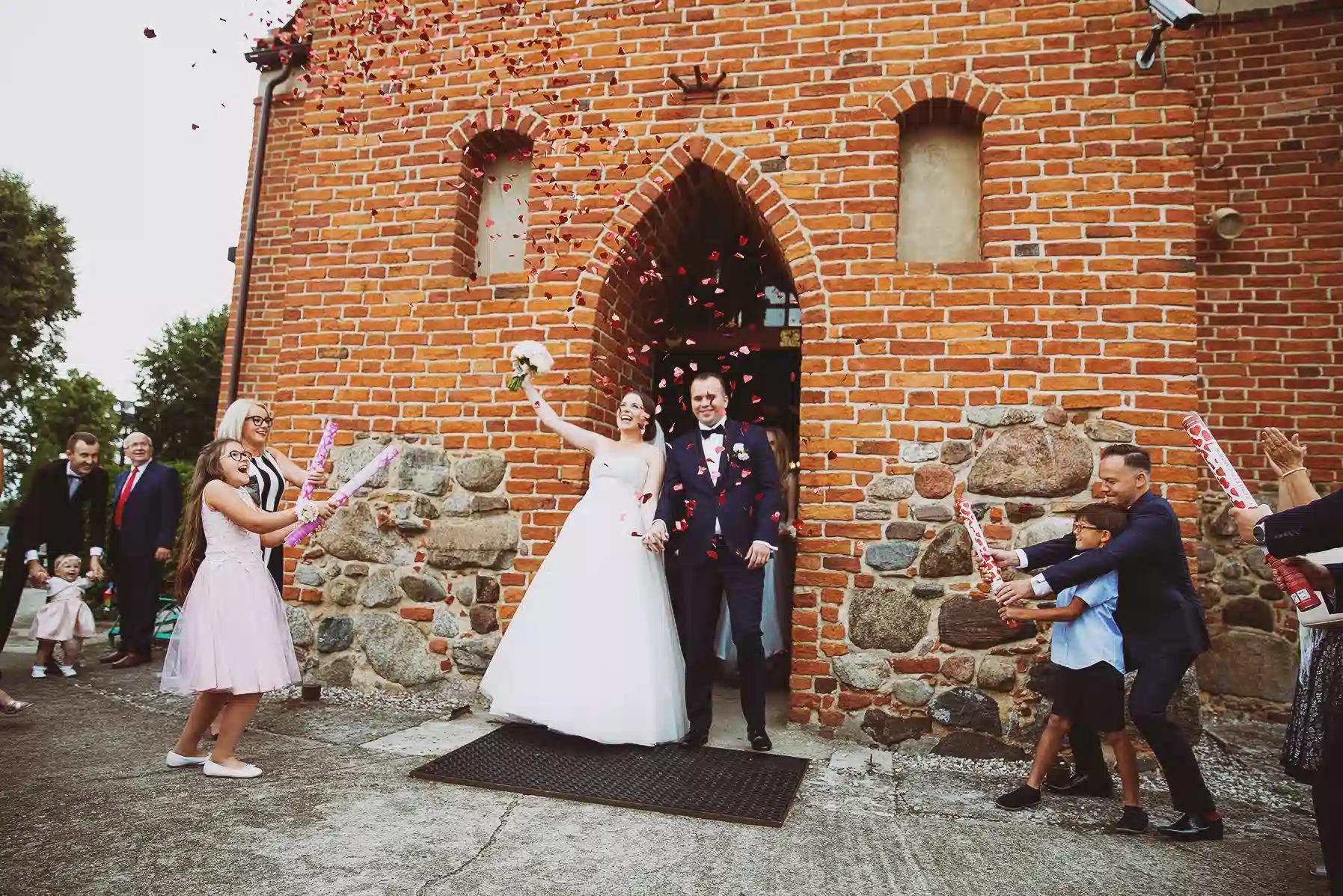 Para młoda wychodzi z kościoła obsypywana konfetti, w tle ceglana fasada i wiwatujący goście z tubami. Kadr z reportażu ślubnego.