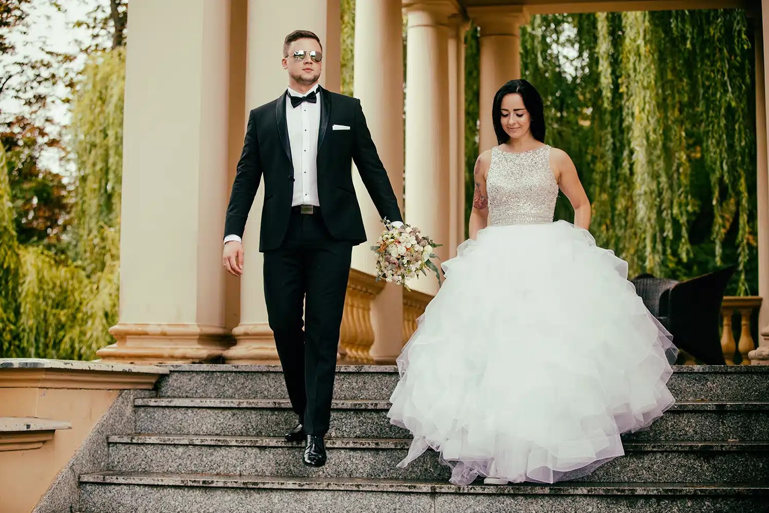 Elegancka para młoda schodzi po schodach, trzymając się za ręce. Pan młody w czarnym smokingu i okularach, panna młoda w białej sukni z bukietem. Kolumnada w tle.