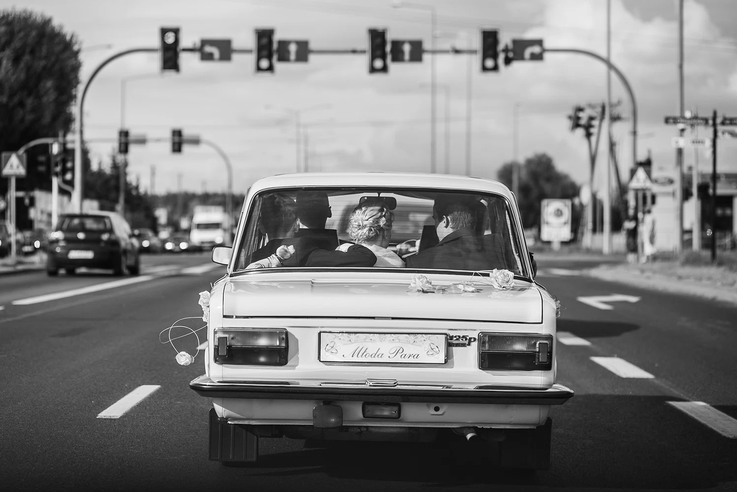Czarno-białe zdjęcie ślubne: Młoda para w białym Fiacie 125p z dekoracją kwiatową i tablicą 'Młoda Para' na tle miejskiej ulicy z sygnalizacją świetlną.