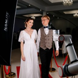 Adamatic - Elegancka para młoda pozuje przed robotem filmowym Glambot na czerwonym dywanie. W tle żyrandole i dekoracje weselne, oświetlenie studyjne.