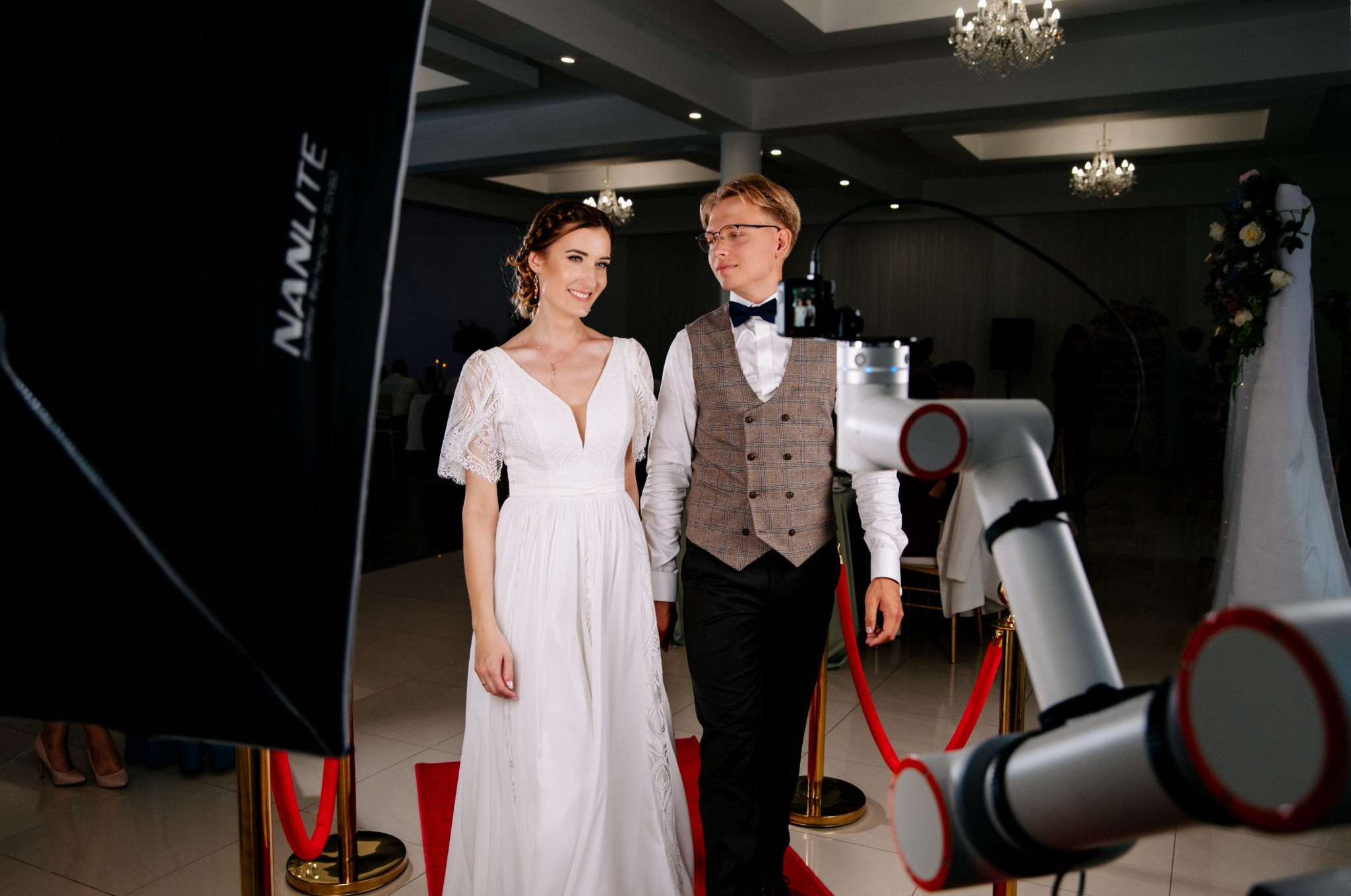 Elegancka para młoda pozuje przed robotem filmowym Glambot na czerwonym dywanie. W tle żyrandole i dekoracje weselne, oświetlenie studyjne.