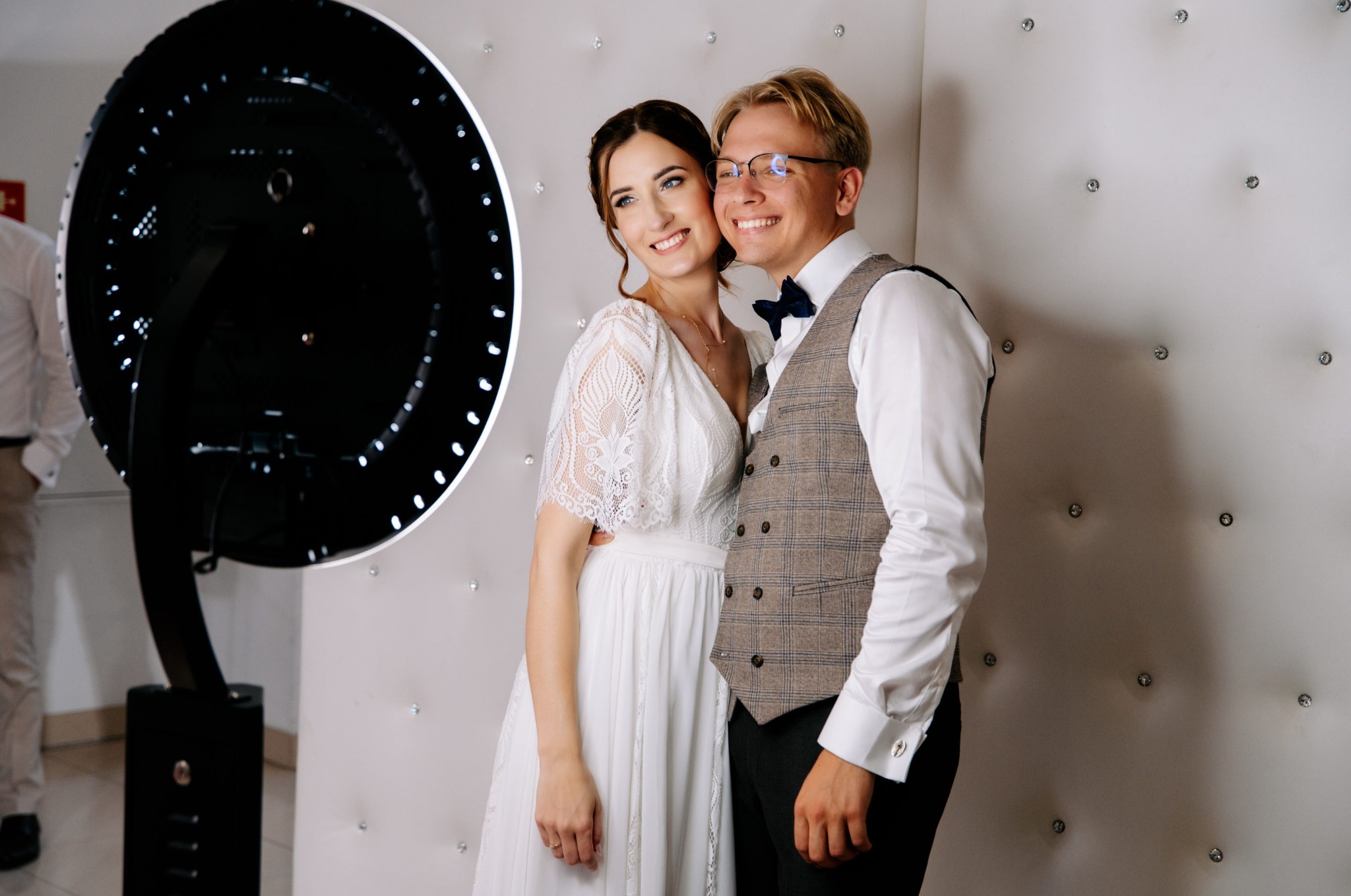 Uśmiechnięta para młoda pozuje przy nowoczesnej fotobudce z oświetleniem LED na tle białej ściany z dekoracyjnymi kryształkami. Pan młody w kamizelce.