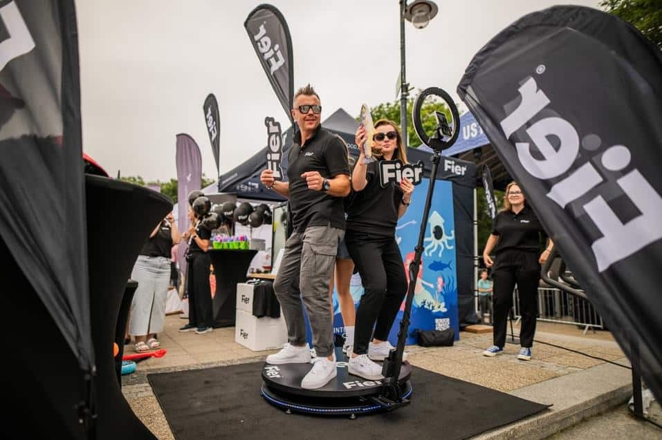 Dwie osoby pozują na platformie fotobudki 360 z rekwizytami. W tle widoczny namiot firmowy, balony i obsługa. Uczestnicy w okularach przeciwsłonecznych i czarnych koszulkach.