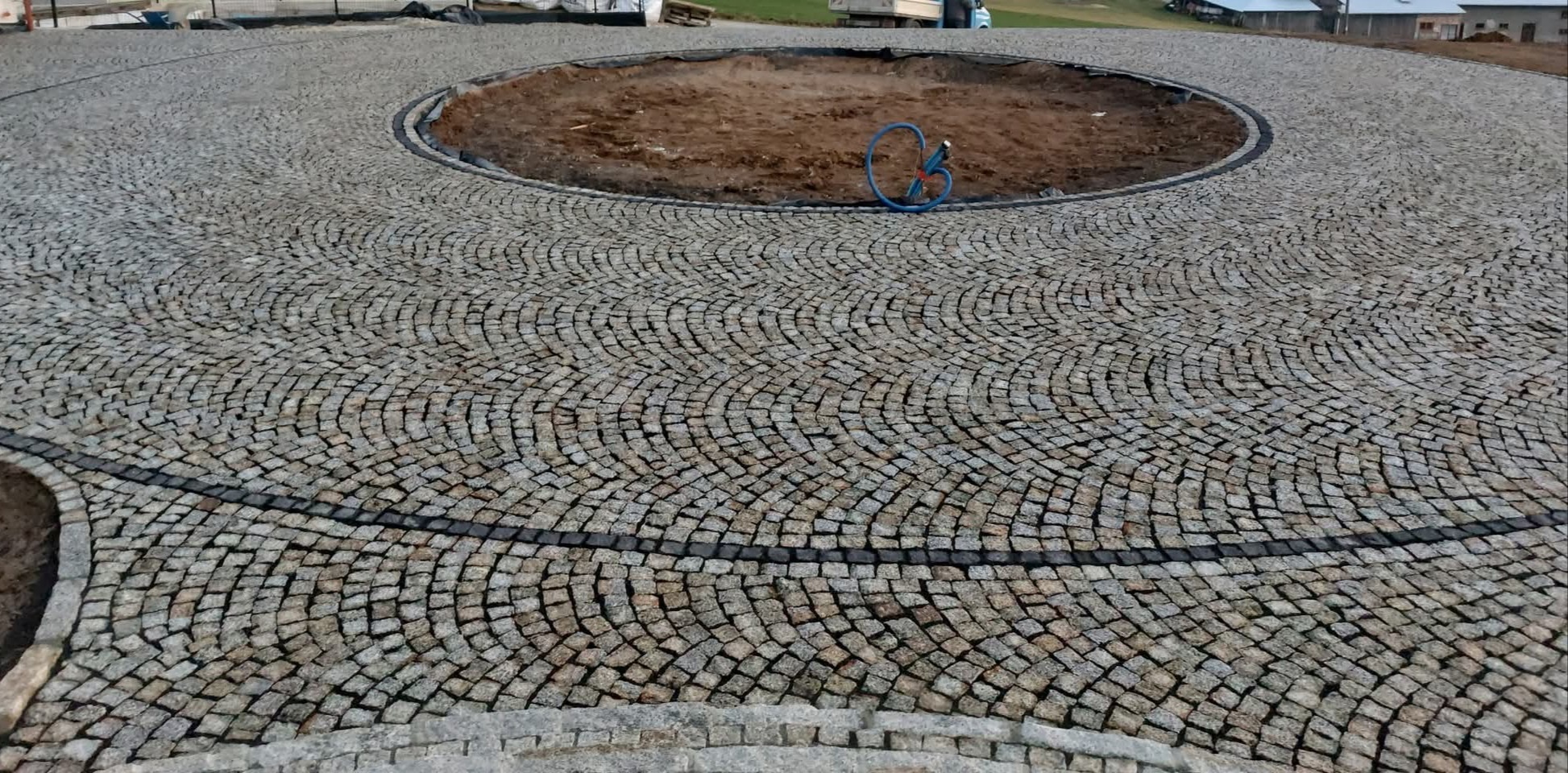 Granitowa nawierzchnia ułożona w okrąg z centralnym klombem w trakcie tworzenia. Widoczne detale wzoru i obrzeża. Na klombie leży niebieski wąż.