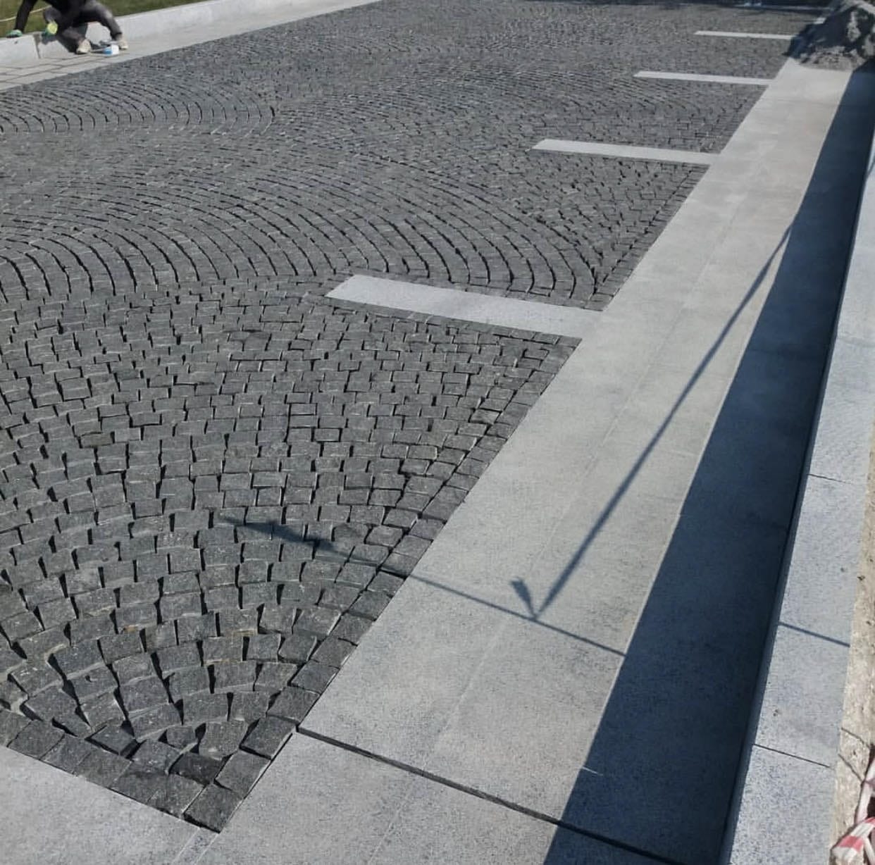 Szara kostka granitowa ułożona w łuki i proste linie, oddzielona jaśniejszymi pasami i obrzeżem. Widoczny fragment osoby w lewym górnym rogu i cień słupa.