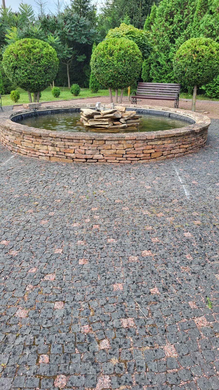 Okrągła fontanna z kamienia na tle brukowanej powierzchni i zieleni parkowej. Widoczne detale układania kostki i naturalne otoczenie.
