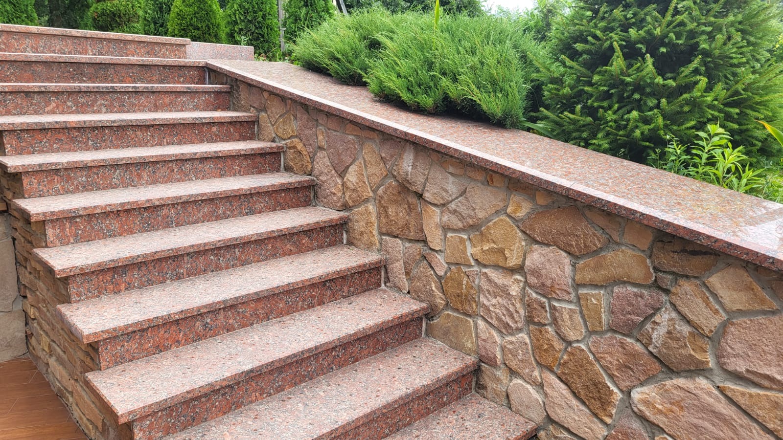 Granitowe schody zewnętrzne z elementami kamiennej elewacji i zieleni w tle, wykonane z czerwonego granitu, prezentują się elegancko i trwale.