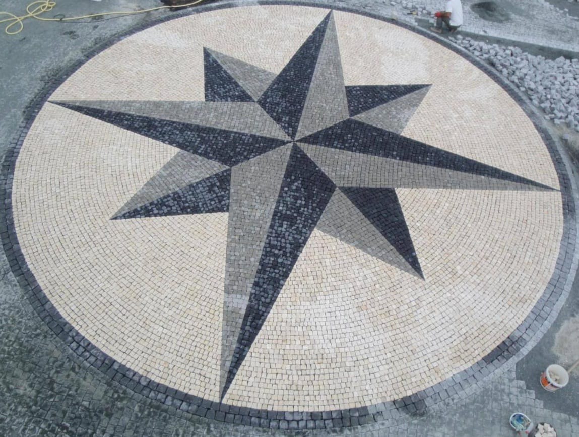 Granitowa mozaika w kształcie gwiazdy wkomponowana w okrągłą nawierzchnię z jasnej kostki, obrzeża z ciemnej kostki. Prace brukarskie w toku, widoczny pracownik i materiały.