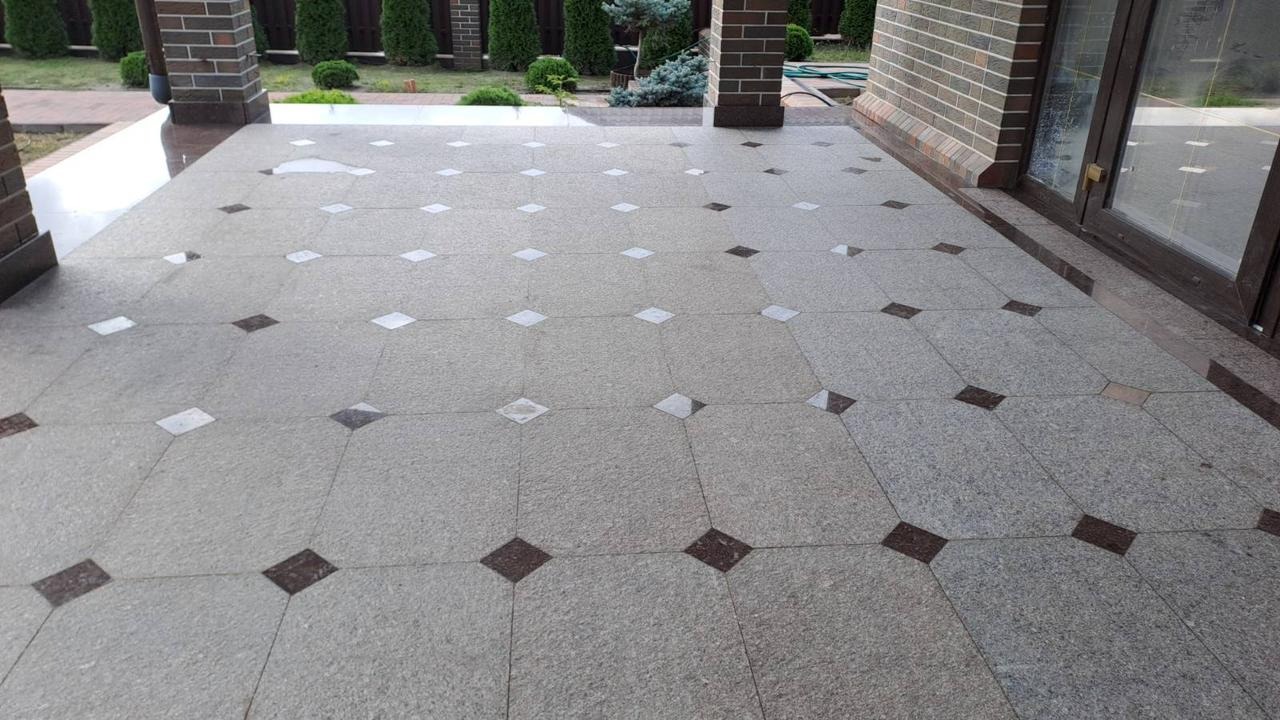 Wykończony taras z szarej kostki granitowej z wstawkami z białych i brązowych kostek w kształcie rombów. Widoczna kolumna i fragment ściany budynku z cegły.