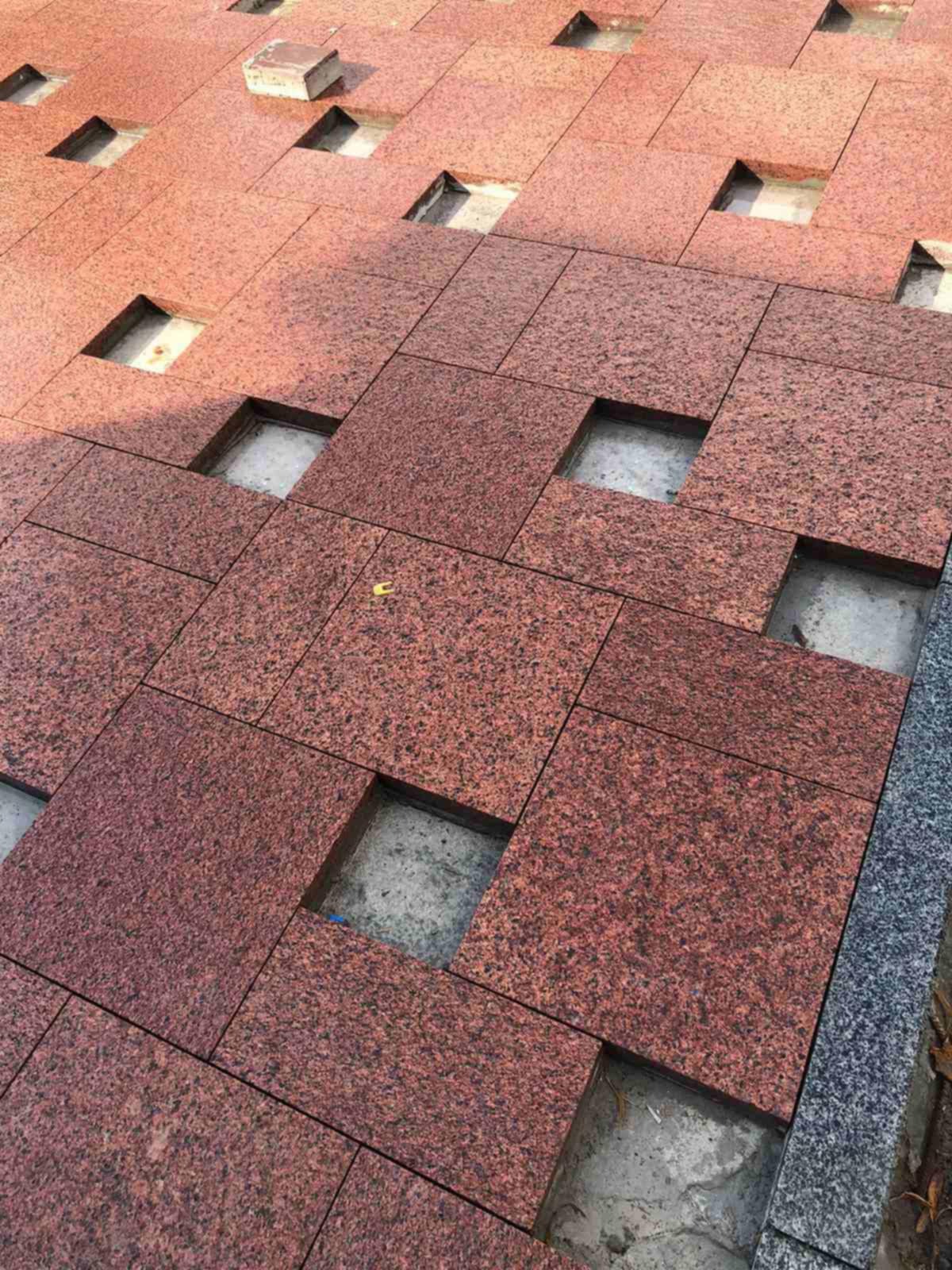 Czerwona kostka granitowa ułożona w kwadraty, część kostek brakuje, odsłaniając podłoże. Widok z góry, z bliska.