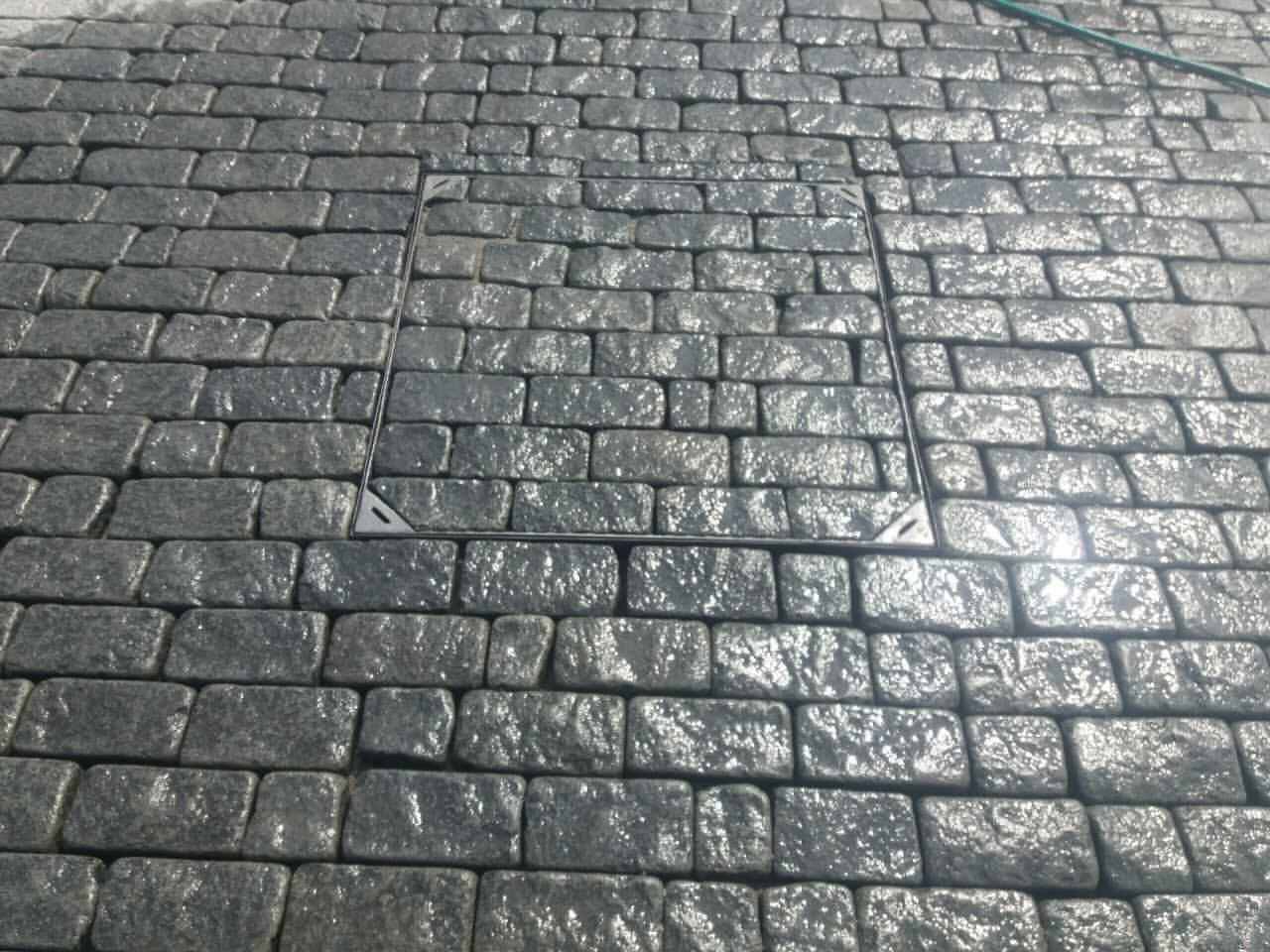 Szara kostka brukowa ułożona w regularny wzór z metalową pokrywą włazu pośrodku. Widoczne odbicia światła na mokrej powierzchni kostki i fragment węża ogrodowego.