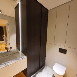 Deineka.pl - Nowoczesna łazienka w trakcie wykańczania: ciemna szafa z lustrem, umywalka z marmurowym blatem, biała toaleta. Podłoga wyłożona marmurowymi płytami. Folie ochronne na podłodze.