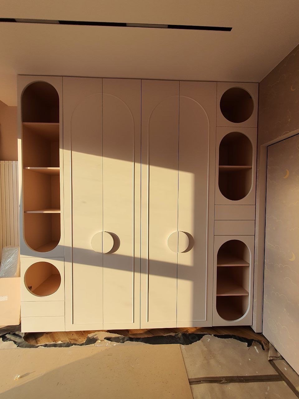 Biała szafa na wymiar z okrągłymi uchwytami i półkami, oświetlona popołudniowym słońcem, w pokoju z tapetą w chmurki. Widoczne niedoskonałości podłogi.