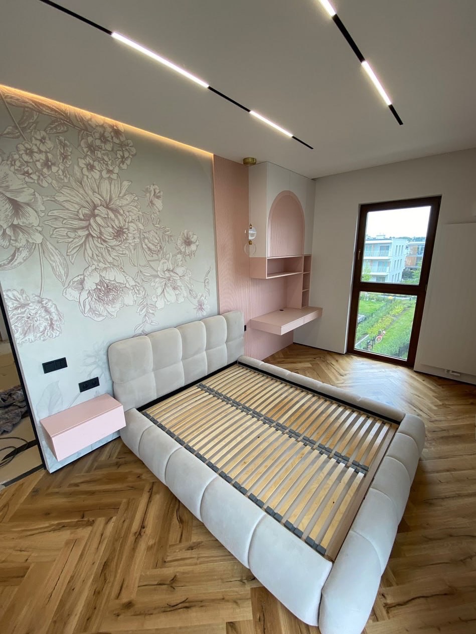 Beżowe łóżko z tapicerowanym zagłówkiem i drewnianym stelażem na parkiecie w jodełkę, z dekoracyjną ścianą w kwiaty i biurkiem w odcieniach różu.