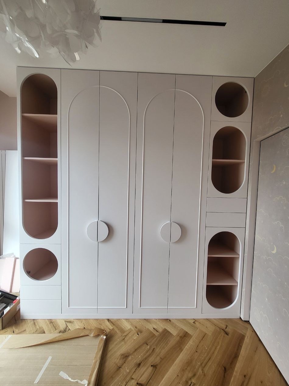 Biała szafa wnękowa z zaokrąglonymi elementami i okrągłymi uchwytami, na parkiecie ułożonym w jodełkę. Widoczne półki z różowym wnętrzem i ozdobna tapeta w tle.