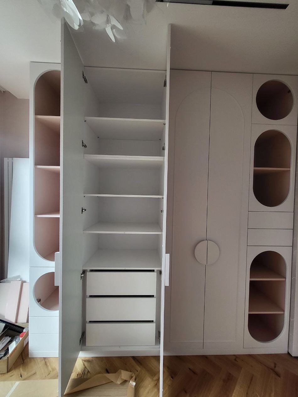 Biała szafa wnękowa z półkami i szufladami, widok z otwartymi drzwiami, nietypowe okrągłe wnęki z półkami po prawej stronie, parkiet na podłodze.
