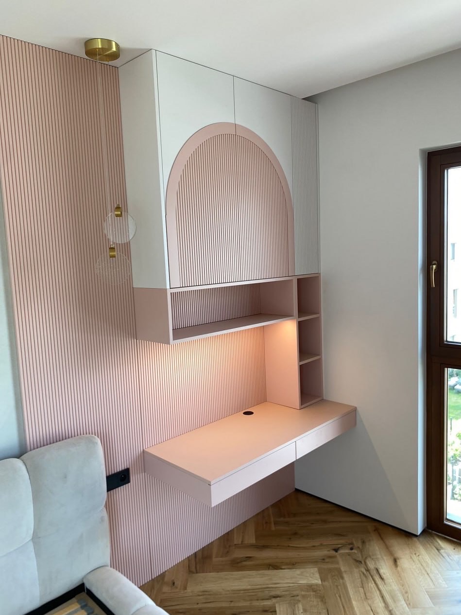 Różowo-białe biurko na wymiar z szafkami wiszącymi, oświetleniem i dekoracyjną ścianą z lameli. Widok na jasne wnętrze z parkietem.