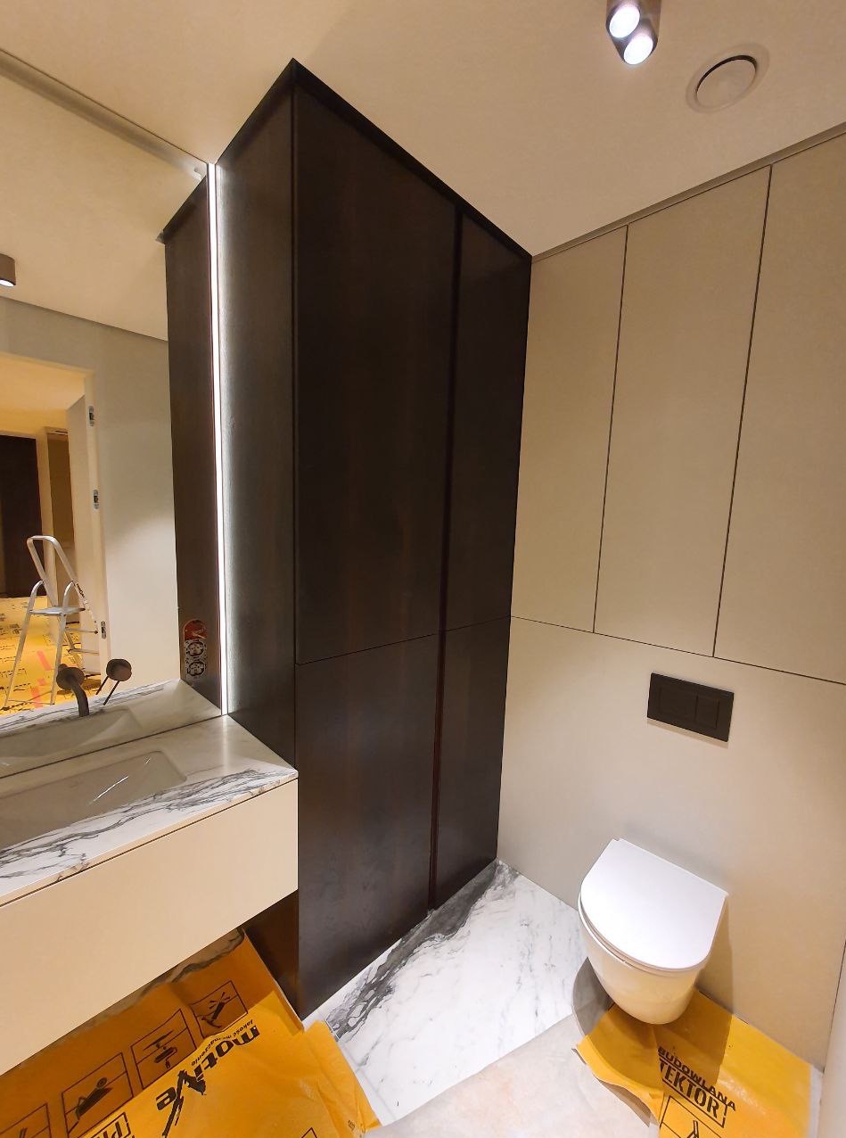 Nowoczesna łazienka w trakcie wykańczania: ciemna szafa z lustrem, umywalka z marmurowym blatem, biała toaleta. Podłoga wyłożona marmurowymi płytami. Folie ochronne na podłodze.