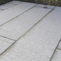 Szary dach z papy termozgrzewalnej, widoczne łączenia i detale wykończenia, fragment zieleni w tle. Ujęcie z góry, ukazujące strukturę i fakturę materiału.