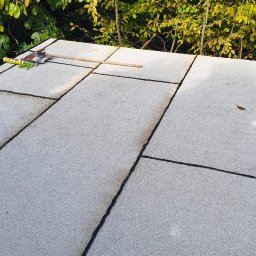 NATH-BUD DANIEL KASTELIK - Płaski dach pokryty papą termozgrzewalną w jasnoszarym kolorze, z widocznymi narzędziami dekarskimi i zielenią w tle. Widok z góry na wykończony dach.