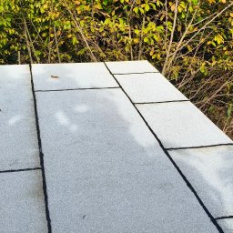 NATH-BUD DANIEL KASTELIK - Szary dach pokryty papą termozgrzewalną na tle zieleni drzew. Widoczne łączenia papy. Na dachu leżą opadłe liście.