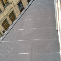 NATH-BUD DANIEL KASTELIK - Szary, płaski dach pokryty papą na budynku wielorodzinnym, widziany z góry pod kątem. W tle zabudowa miejska z oknami i rynnami. Widoczne łączenia papy.