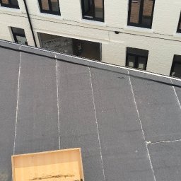 NATH-BUD DANIEL KASTELIK - Czarny dach z papy termozgrzewalnej, na którym stoi drabina z drewnianą skrzynią, biały budynek w tle. Widok z góry, ostre światło.