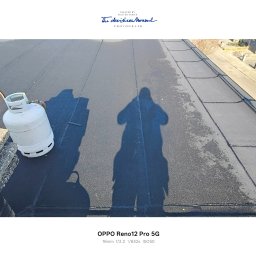 NATH-BUD DANIEL KASTELIK - Czarny, płaski dach po renowacji z widocznym cieniem osoby i butlą gazową. W tle budynki mieszkalne. Widok z góry, ostre światło słoneczne.