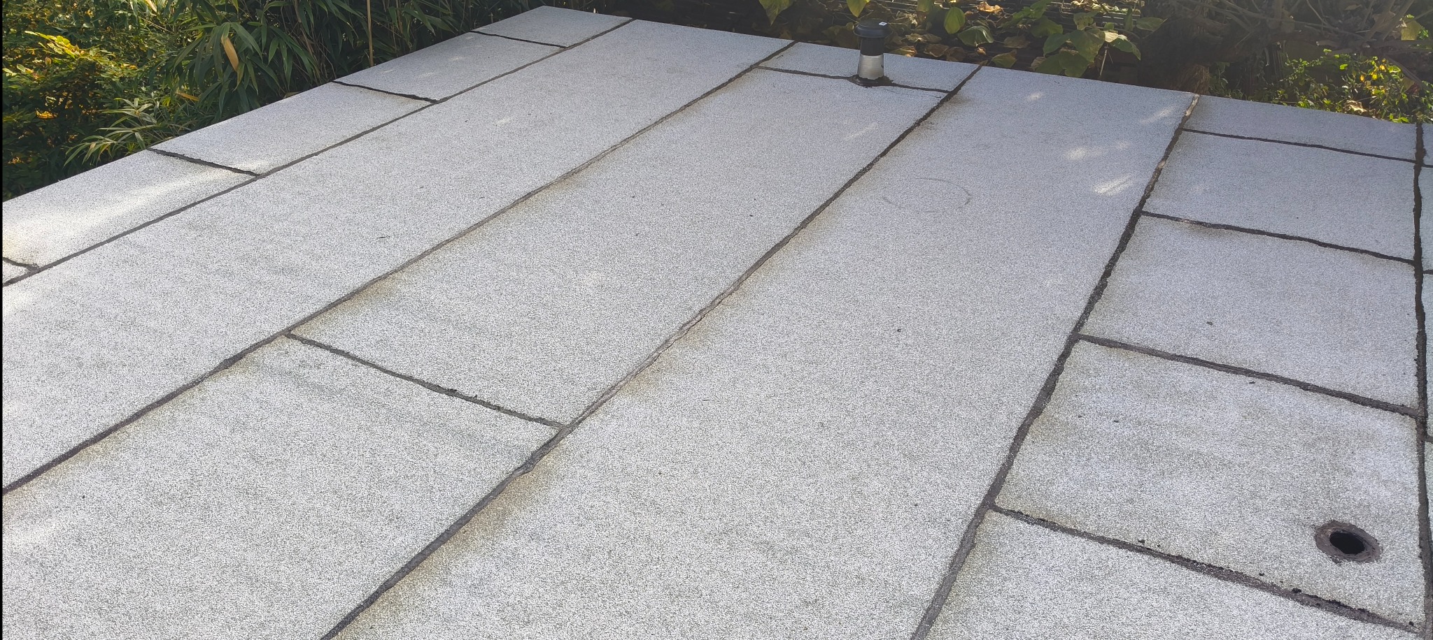 Szary dach z papy termozgrzewalnej, widoczne łączenia i detale wykończenia, fragment zieleni w tle. Ujęcie z góry, ukazujące strukturę i fakturę materiału.