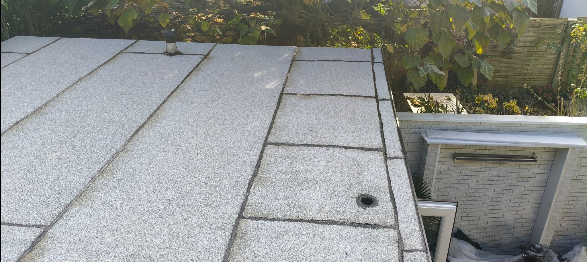 Szary, płaski dach pokryty płytami bitumicznymi, widok z góry, z fragmentem rynny i zieleni w tle.