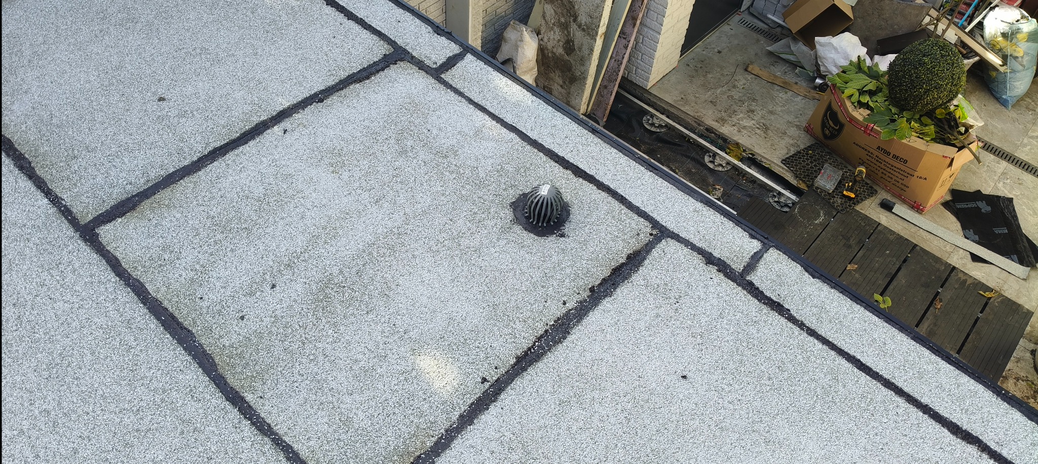 Płaski dach pokryty szarą papą z widocznym wpustem dachowym. Papa podzielona na kwadraty z czarnym uszczelnieniem. Fragment otoczenia dachu.