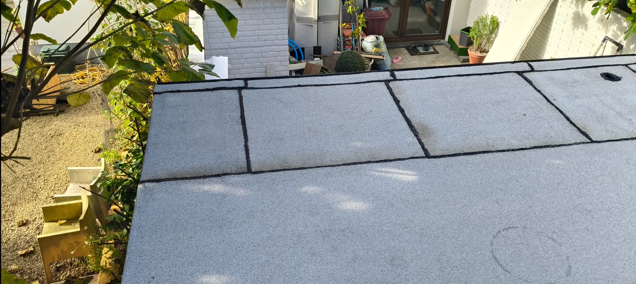 Wykończony dach płaski z papy termozgrzewalnej, widok z góry, z fragmentem roślinności w tle. Szare płyty papy z czarnymi łączeniami.