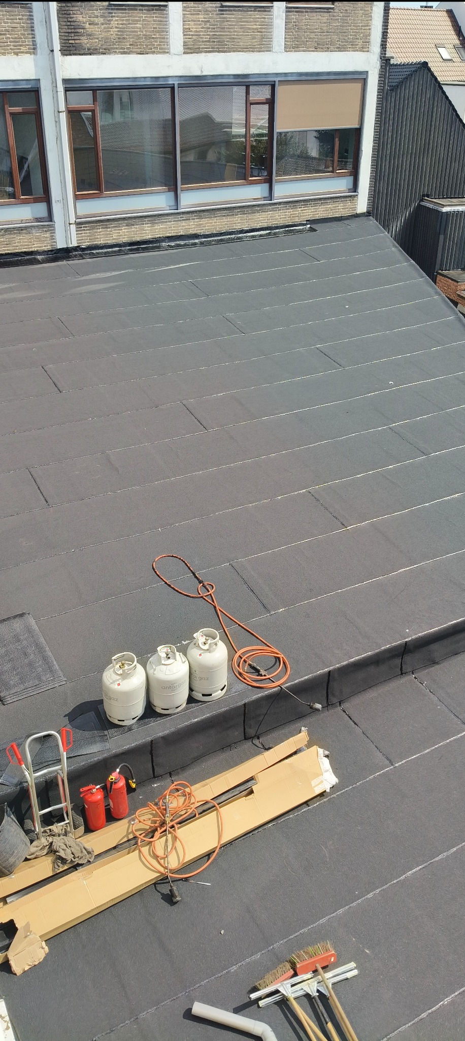 Czarny, płaski dach z papy termozgrzewalnej, na którym leżą narzędzia dekarskie: butle z gazem, palnik, szczotki i drabinka. Widok z góry na budynek.
