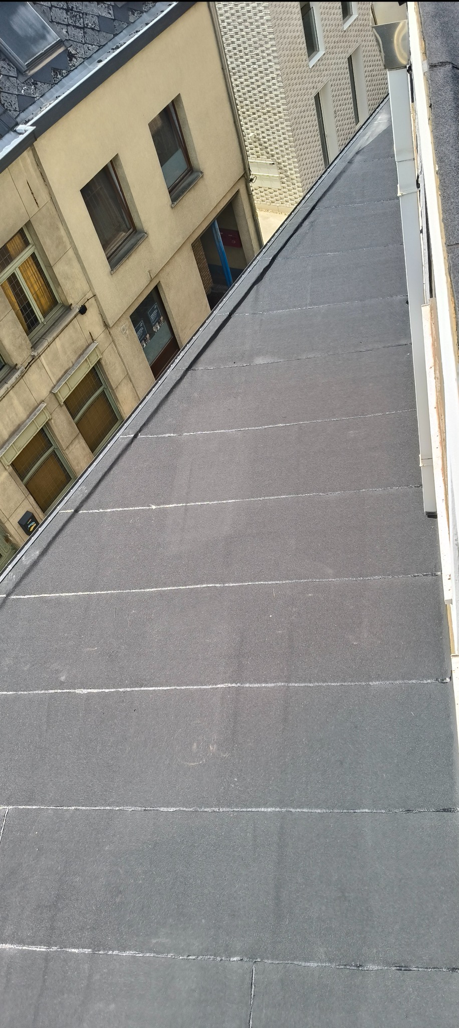 Szary, płaski dach pokryty papą na budynku wielorodzinnym, widziany z góry pod kątem. W tle zabudowa miejska z oknami i rynnami. Widoczne łączenia papy.