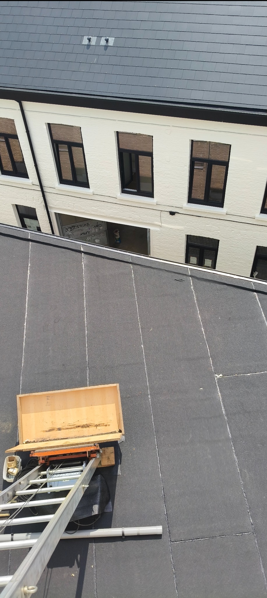 Czarny dach z papy termozgrzewalnej, na którym stoi drabina z drewnianą skrzynią, biały budynek w tle. Widok z góry, ostre światło.