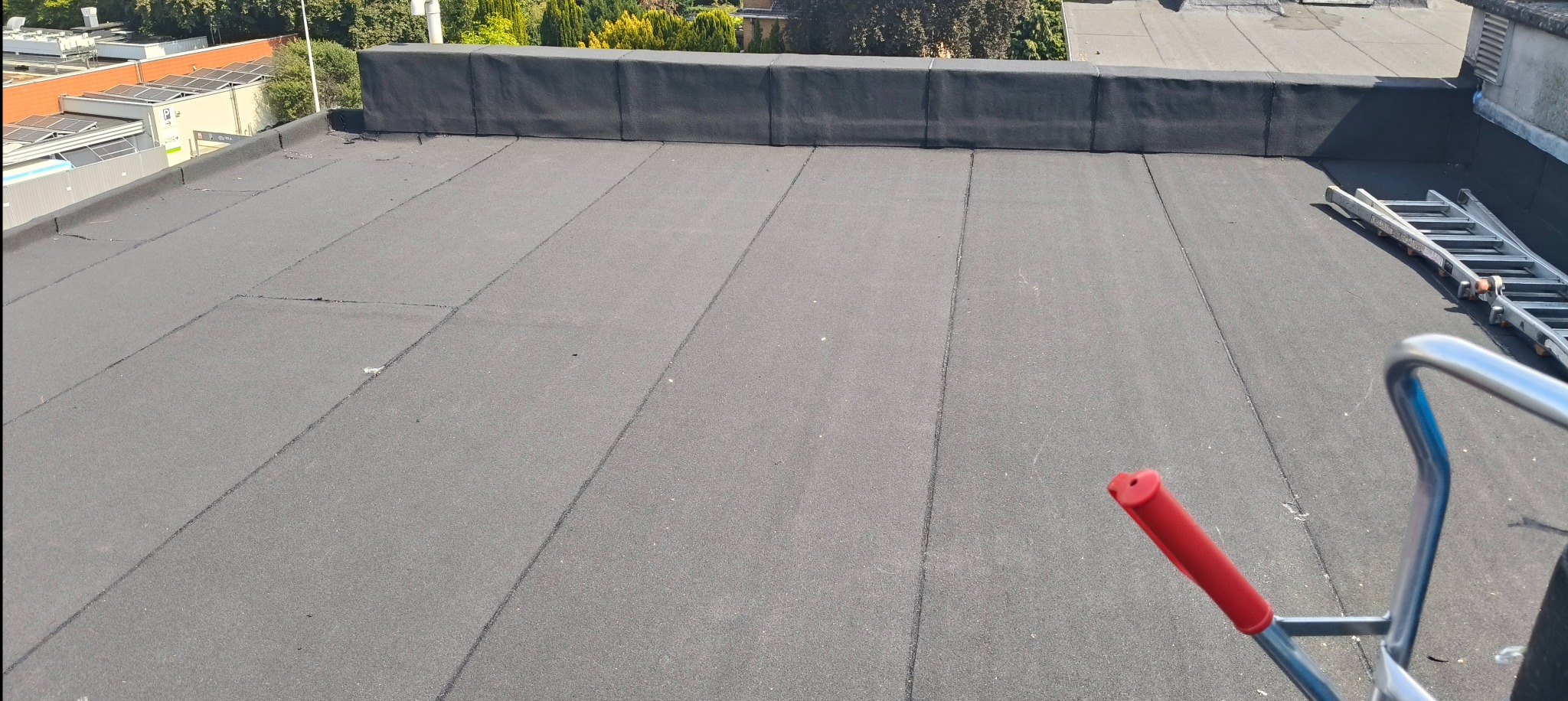 Czarny, płaski dach pokryty papą z widocznymi łączeniami, fragment drabiny dekarskiej, w tle budynki i drzewa w słoneczny dzień.