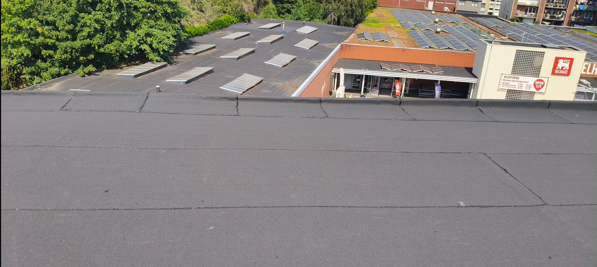 Widok z góry na płaski dach pokryty papą, z widocznymi łączeniami i sąsiednimi budynkami z panelami słonecznymi i świetlikami dachowymi w tle.