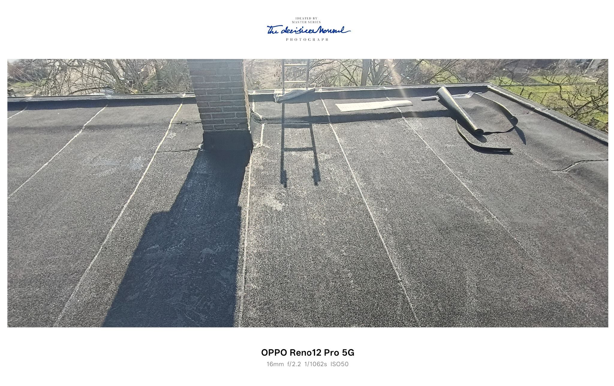 Czarny, płaski dach pokryty papą z widocznym kominem z cegły, fragmentem drabiny i rolką papy. Widoczne łączenia papy. Słoneczny dzień.