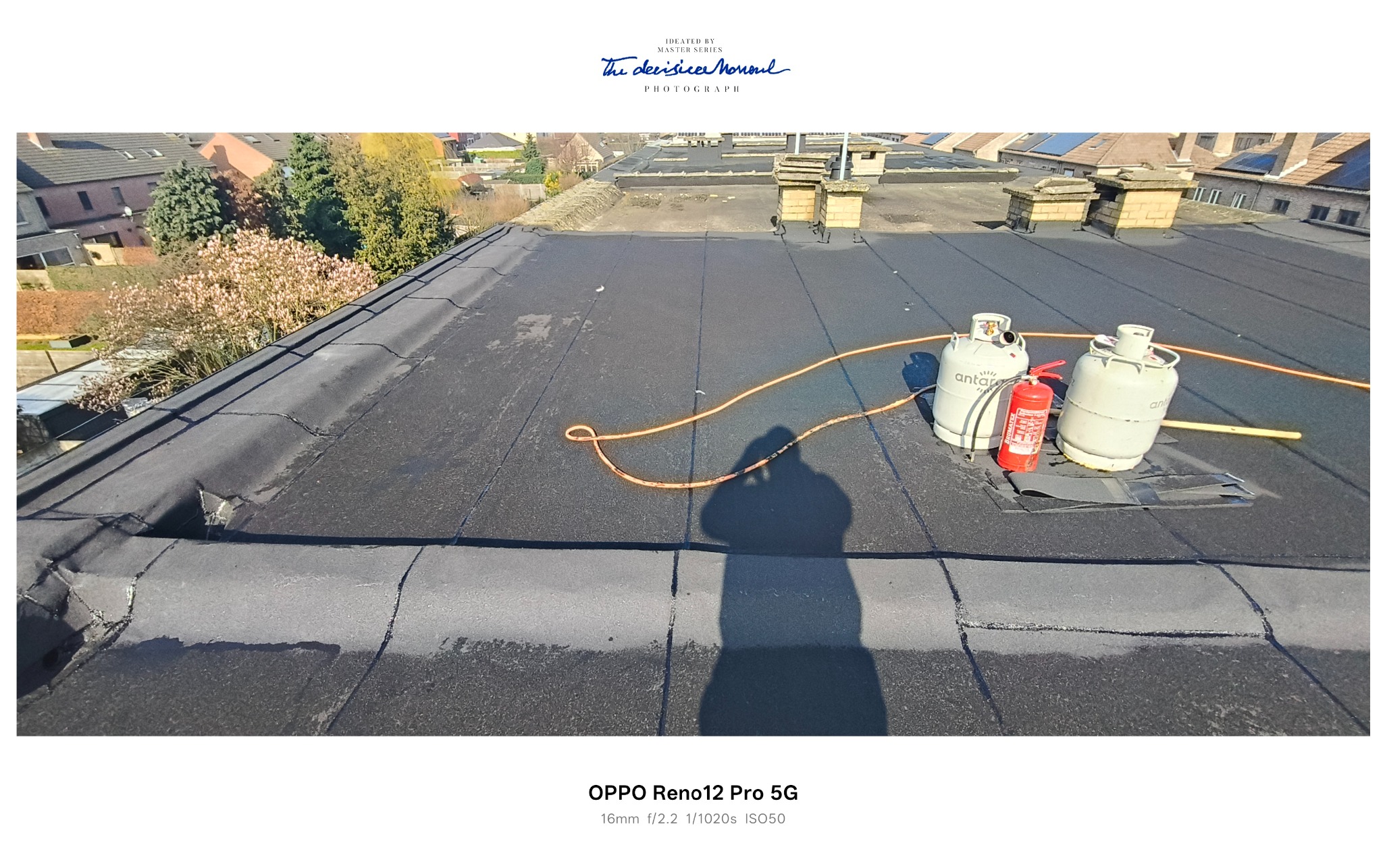 Czarny, płaski dach pokryty papą termozgrzewalną z widocznymi butlami gazowymi, wężem i gaśnicą. W tle budynki mieszkalne i drzewa. Widoczny cień osoby robiącej zdjęcie.
