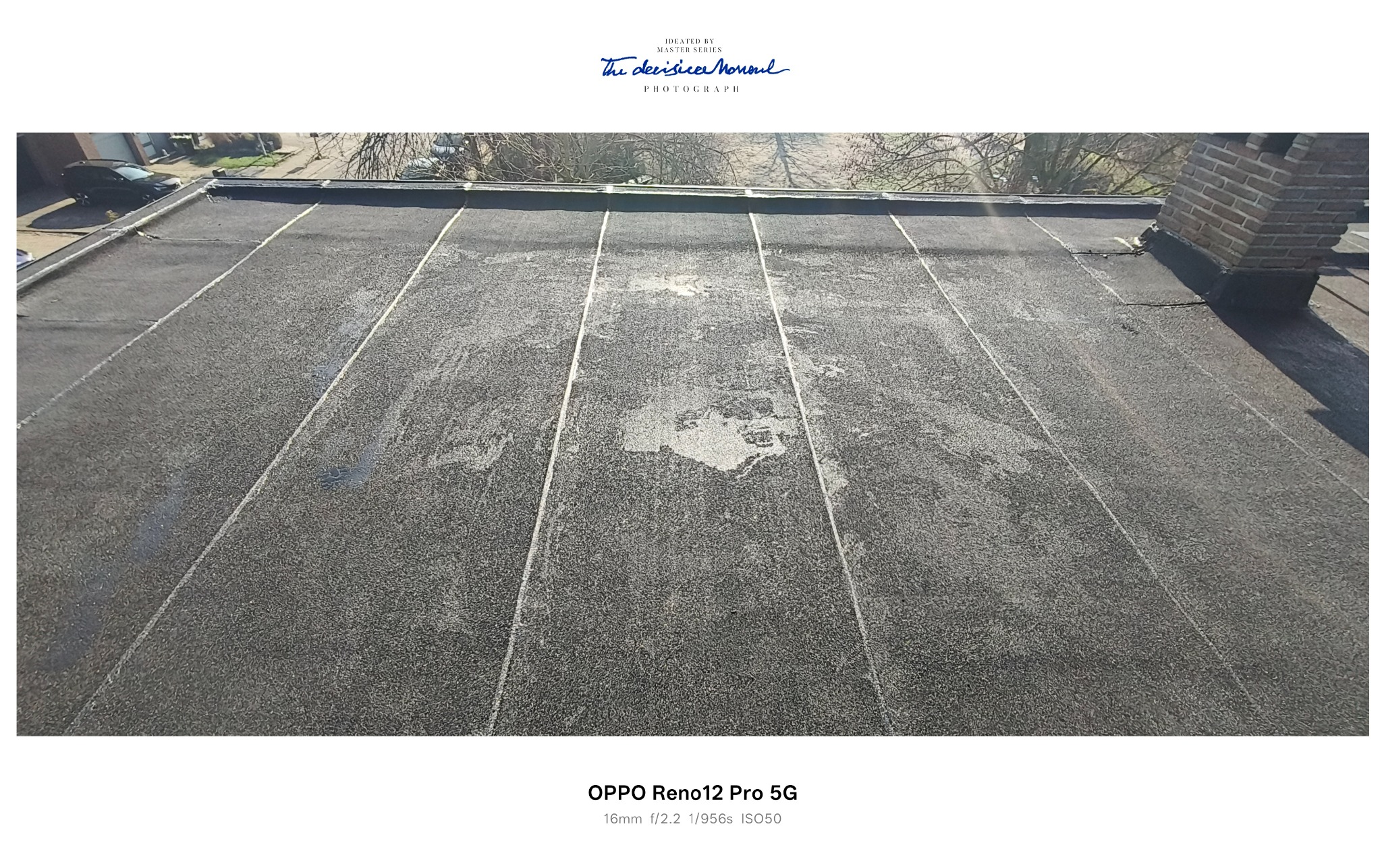 Płaski dach pokryty papą z widocznymi łączeniami i śladami użytkowania, komin w prawym górnym rogu, w tle drzewa i budynki. Widok z góry.